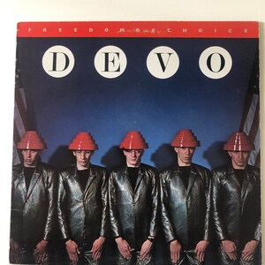 May include: Album cover for Devo's 'Freedom of Choice'. The image shows the band members in silver jumpsuits and red hats, with the word 'DEVO' in white circles. The background features an American flag and the words 'FREEDOM CHOICE'.