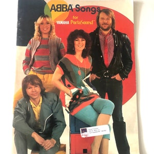 May include: A vintage ABBA songbook for Yamaha PortaSound. The cover shows a photograph of the four ABBA members. The background is red with a yellow, green, and orange circle. The band members are wearing 1970s style clothing.