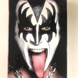 May include: A close-up of a person with white face paint and black markings around the eyes, with their tongue extended. The text "KISS AND MAKE-UP - GENE SIMMONS" is at the bottom. The person is wearing a silver chain.