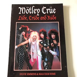 May include: A black hardcover book titled "Mötley Crüe: Lüde, Crüde and Rüde" with a photograph of the band. The cover has red and white text. The authors' names, Sylvie Simmons & Malcolm Dome, are printed at the bottom of the book.