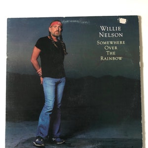 May include: Album cover of Willie Nelson, wearing a black top, blue jeans, and a red bandana. The album title, "Somewhere Over The Rainbow," is in white text against a dark backdrop with a mountain range in the distance.