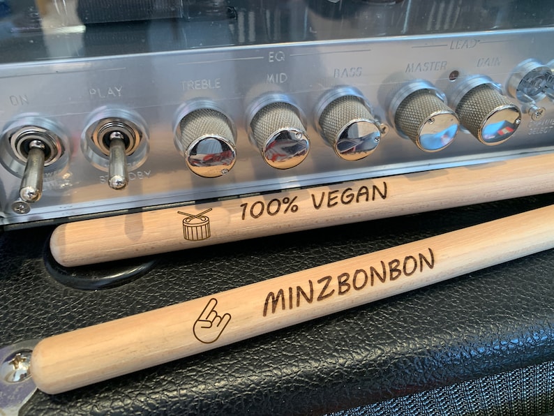 May include: Two wooden drumsticks with engraved text. The top drumstick reads "100% VEGAN" and has a small drum symbol. The bottom drumstick reads "MINZBONBON" and has a rock and roll hand symbol.