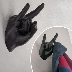 May include: Black metal hand shaped wall hook with two fingers pointing up in a rock and roll hand gesture. A jacket is hanging on the hook.