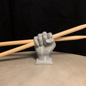 May include: A gray 3D printed hand holding two wooden drumsticks. The hand is on a gray base.