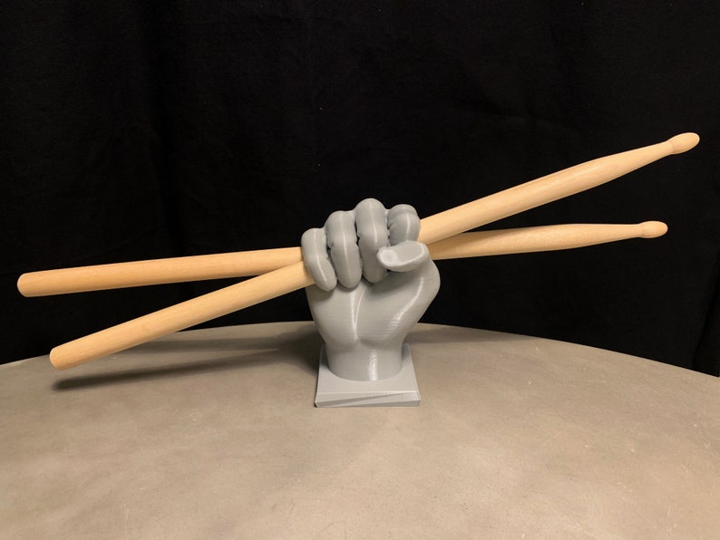 May include: A gray 3D printed hand holding two wooden drumsticks. The hand is on a gray base.