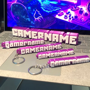 Könnte beinhalten: Fünf violette und weiße 3D-gedruckte Schlüsselanhänger mit dem Wort "gamername" darauf. Die Schlüsselanhänger liegen auf einer Laptop-Tastatur.