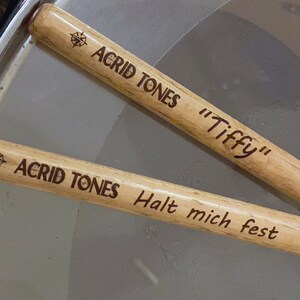 May include: Two wooden drumsticks with the text "Acrid Tones" and a compass symbol on each. One drumstick also has the text "Tiffy" and the other has the text "Halt mich fest".