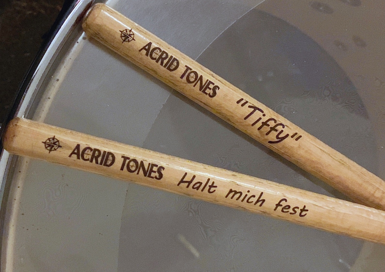 May include: Two wooden drumsticks with the text "Acrid Tones" and a compass symbol on each. One drumstick also has the text "Tiffy" and the other has the text "Halt mich fest".