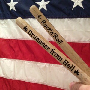 May include: Two wooden drumsticks with the text "Rock'n'Roll" and "Drummer from Hell" engraved on them. The drumsticks are placed on top of an American flag.