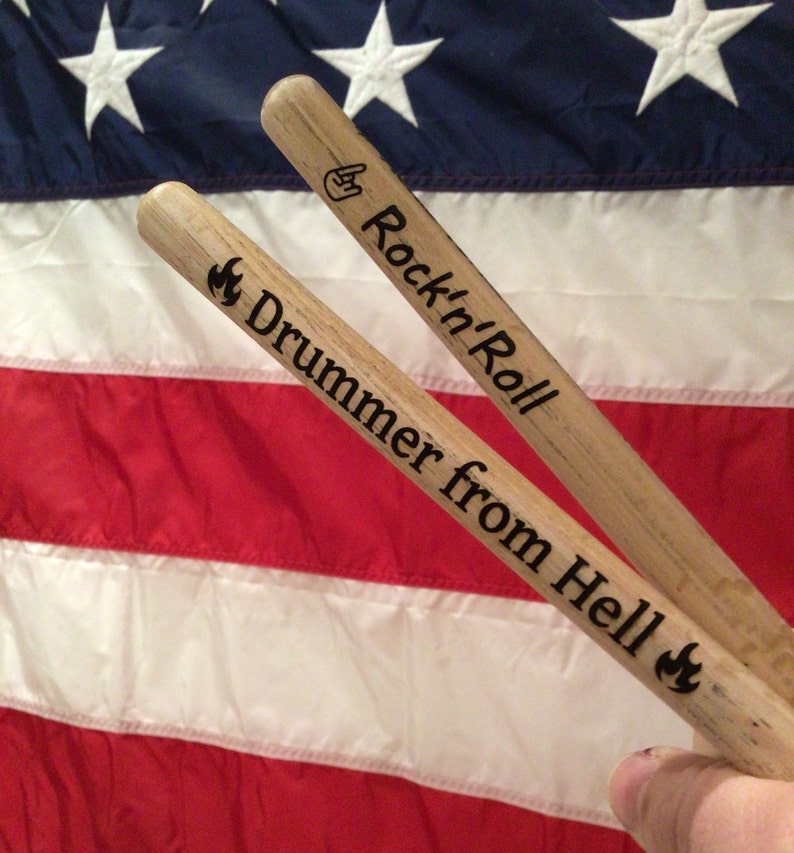 May include: Two wooden drumsticks with the text "Rock'n'Roll" and "Drummer from Hell" engraved on them. The drumsticks are placed on top of an American flag.