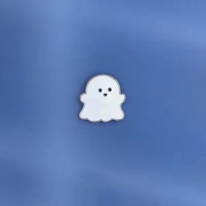 May include: A white enamel pin shaped like a ghost with a smiling face. The ghost has a silver outline.