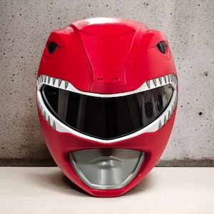 May include: A red plastic Power Rangers helmet with a white visor and a silver mouth guard. The helmet has a dinosaur-like design with white teeth and black eyes.