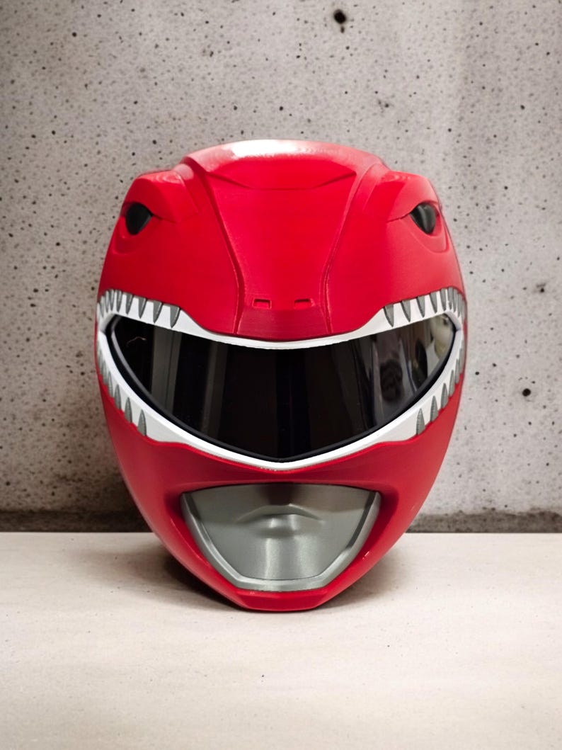 May include: A red plastic Power Rangers helmet with a white visor and a silver mouth guard. The helmet has a dinosaur-like design with white teeth and black eyes.