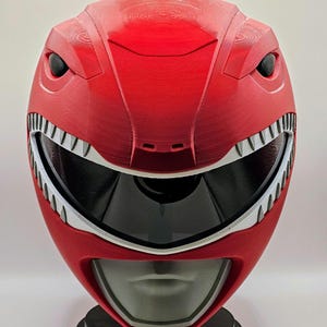 May include: A red and white plastic helmet with a visor and a dinosaur-like design. The helmet has a white stripe around the visor and white teeth around the mouth.