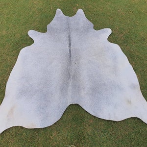 May include: A light gray cowhide rug with a natural, slightly textured surface. The rug is laid out on a green grassy surface.