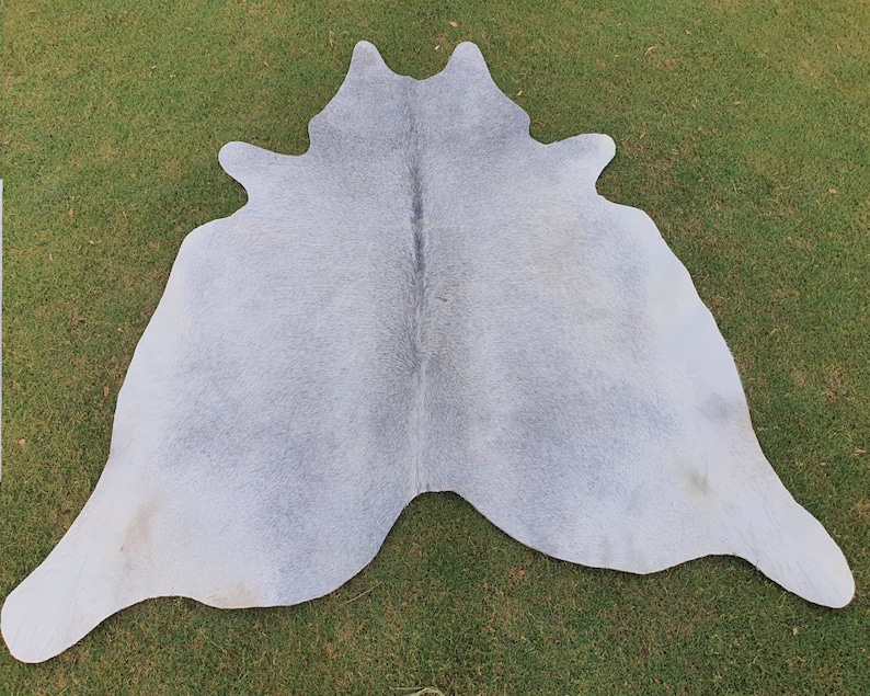 May include: A light gray cowhide rug with a natural, slightly textured surface. The rug is laid out on a green grassy surface.