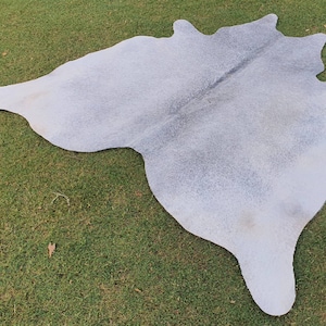 May include: A white cowhide rug with a light gray color variation. The rug is laid out on a green grassy surface.