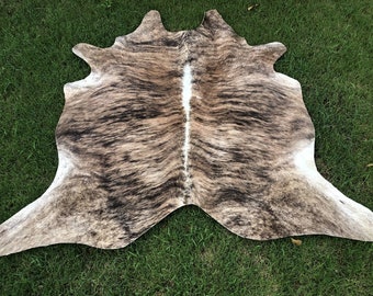 May include: A brown and white cowhide rug with a natural, spotted pattern. The rug is laid out on a green grassy surface.
