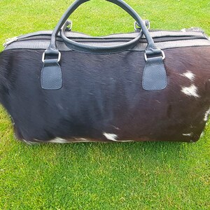 May include: A black and white cowhide leather duffel bag with black leather handles and a zip closure.