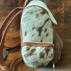 Puede incluir: Una mochila de cuero marr&oacute;n con un panel frontal de piel de vaca blanca y marr&oacute;n. La mochila tiene dos correas ajustables y una cremallera.