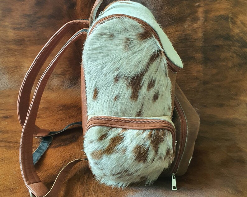 Puede incluir: Una mochila de cuero marr&oacute;n con un panel frontal de piel de vaca blanca y marr&oacute;n. La mochila tiene dos correas ajustables y una cremallera.
