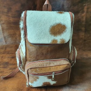 Puede incluir: Una mochila de cuero de vaca marr&oacute;n y blanco con un bolsillo frontal grande y un bolsillo m&aacute;s peque&ntilde;o debajo. La mochila tiene una correa de cuero marr&oacute;n y un cierre de cremallera.