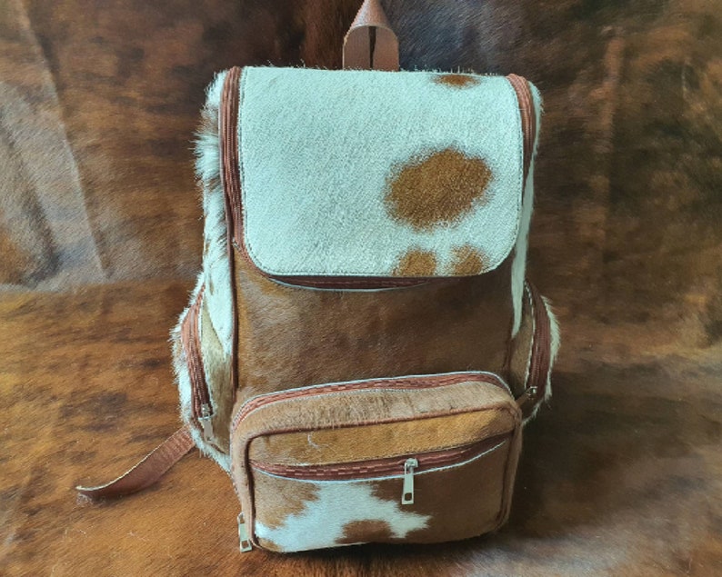 Puede incluir: Una mochila de cuero de vaca marr&oacute;n y blanco con un bolsillo frontal grande y un bolsillo m&aacute;s peque&ntilde;o debajo. La mochila tiene una correa de cuero marr&oacute;n y un cierre de cremallera.