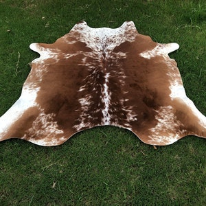 May include: Brown and white cowhide rug with a spotted pattern. The rug is laid out on a green grassy surface.