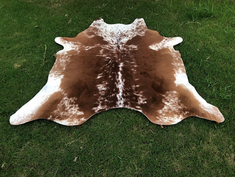 May include: Brown and white cowhide rug with a spotted pattern. The rug is laid out on a green grassy surface.