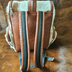 Puede incluir: Mochila de cuero marr&oacute;n con correas negras y un patr&oacute;n de piel de vaca blanca y marr&oacute;n.