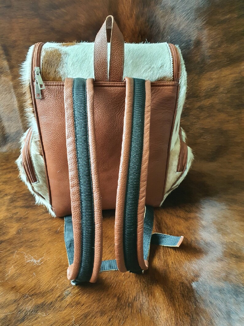 Puede incluir: Mochila de cuero marr&oacute;n con correas negras y un patr&oacute;n de piel de vaca blanca y marr&oacute;n.
