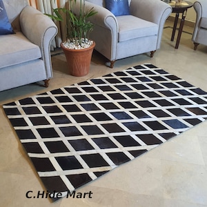 Puede incluir: Alfombra de piel de vaca negra y blanca con un patrón de diamantes. La alfombra está colocada sobre un suelo beige claro delante de dos sillones.