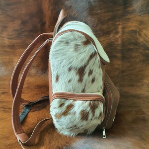 Puede incluir: Una mochila de piel de vaca marr&oacute;n y blanca con cierre de cremallera. La mochila tiene una correa de cuero marr&oacute;n y un asa de cuero marr&oacute;n.