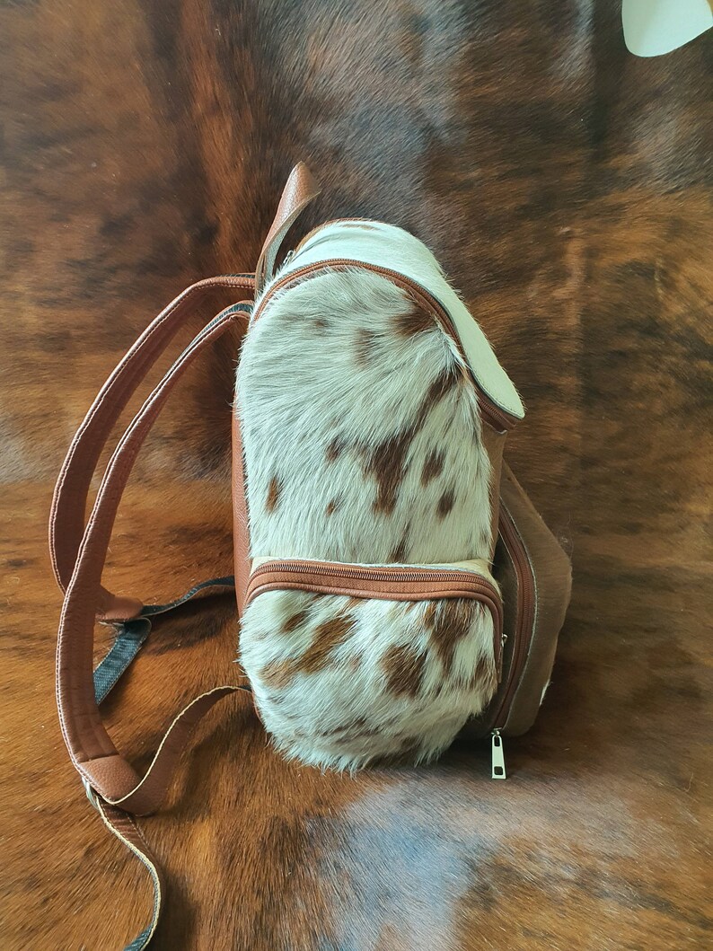 Puede incluir: Una mochila de piel de vaca marr&oacute;n y blanca con cierre de cremallera. La mochila tiene una correa de cuero marr&oacute;n y un asa de cuero marr&oacute;n.