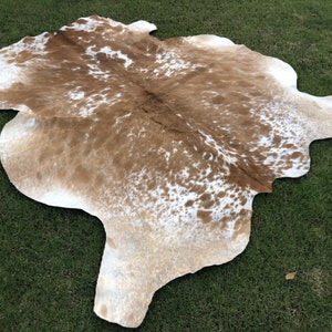 May include: A brown and white cowhide rug with a natural, spotted pattern. The rug is laid out on a green grassy surface.