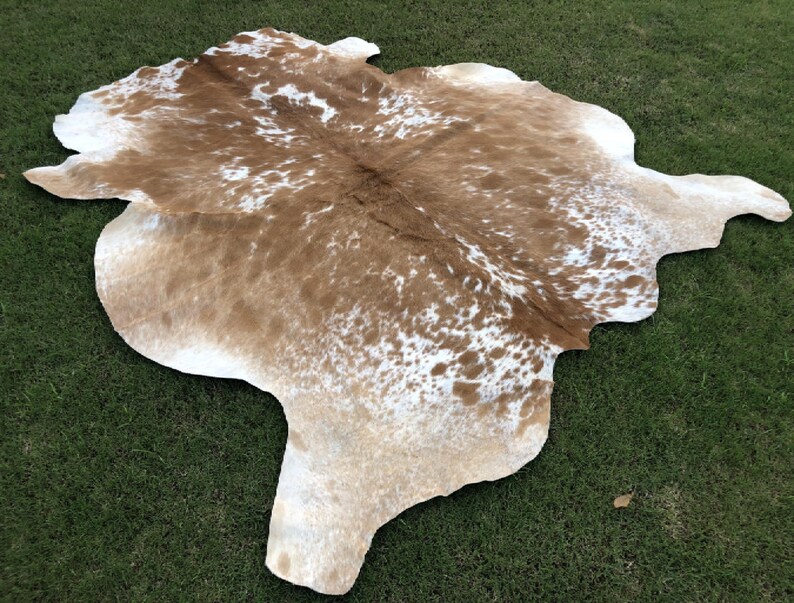 May include: A brown and white cowhide rug with a natural, spotted pattern. The rug is laid out on a green grassy surface.