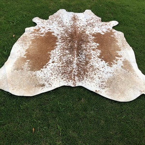 May include: A brown and white cowhide rug with a spotted pattern. The rug is laid out on a green grassy surface.
