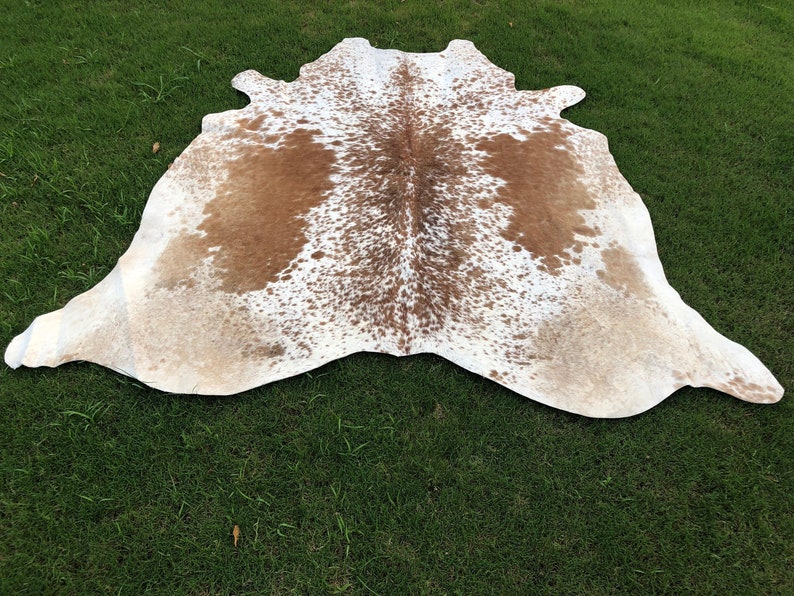 May include: A brown and white cowhide rug with a spotted pattern. The rug is laid out on a green grassy surface.