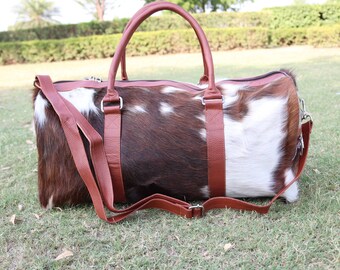 May include: A brown and white cowhide leather duffel bag with brown leather straps and handles.