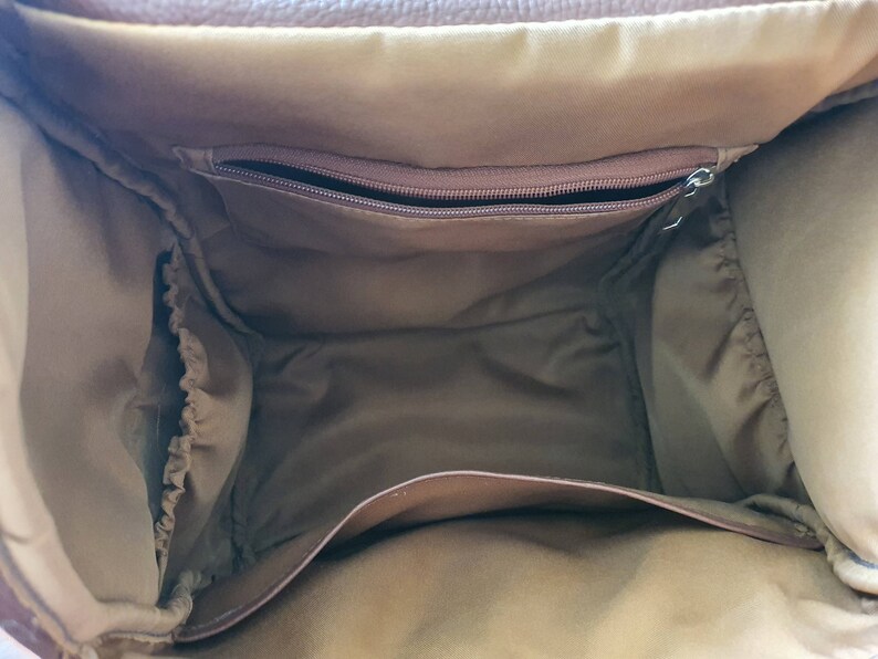 Puede incluir: El interior de una bolsa de pa&ntilde;ales de cuero marr&oacute;n con un bolsillo con cremallera y bucles el&aacute;sticos.