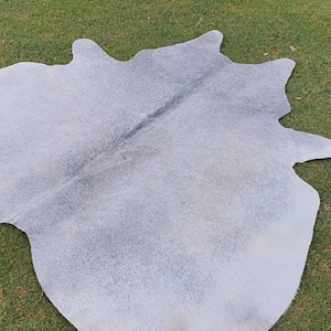 May include: A light gray cowhide rug with a natural, soft texture. The rug is laid out on a green grassy surface.