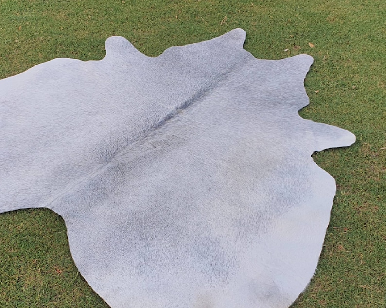 May include: A light gray cowhide rug with a natural, soft texture. The rug is laid out on a green grassy surface.
