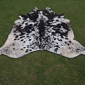May include: Black and white cowhide rug with a spotted pattern. The rug is laid out on a green grassy surface.
