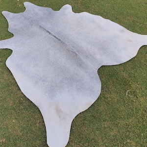 May include: A light gray cowhide rug with a natural, irregular shape. The rug has a soft, fuzzy texture and is laid out on a green grassy surface.