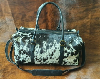 Könnte beinhalten: Schwarze und weiße Kuhfell-Reisetasche mit schwarzen Ledergriffen und einem Schultergurt. Die Tasche hat einen Reißverschluss und Messing-Schnallen.