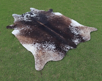 May include: A cowhide rug with a natural pattern of brown, white, and black spots. The rug is irregularly shaped and lies on a green grassy surface. The hide has a soft, textured appearance.