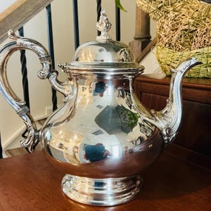 May include: A polished silver teapot with a rounded body, a curved handle, and a decorative finial on the lid. The surface is highly reflective, showing distorted reflections of the surrounding room. It features a wide base and a spout.