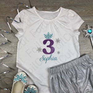 Könnte beinhalten: Ein weißes T-Shirt mit einer lilafarbenen Zahl 3, einer Schneeflockenkrone und dem Namen Sophia in silbernem Glitzer. Das Shirt wird mit silbernen Glitzer-Shorts, einem silbernen Zauberstab mit einer blauen Schneeflocke und einem blauen Herzen sowie weißen Schuhen mit blauen Schneeflocken-Verzierungen kombiniert.