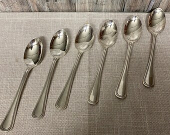 May include: Six silver spoons with beaded edges. The spoons are arranged in a row on a light brown fabric surface.