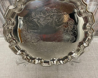 May include: A silver serving tray with a scalloped edge and an ornate engraved design. The tray is on a stand.
